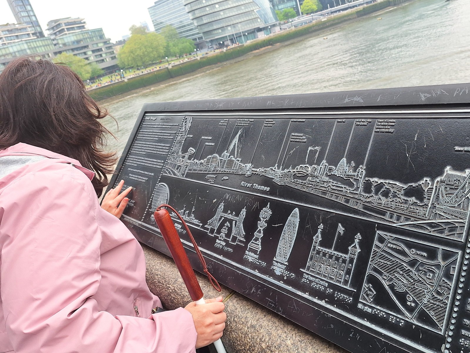 Елена проследява с пръсти тактилен модел на открито в облачен ден, бастунът ѝ е подпрян на лакътя.