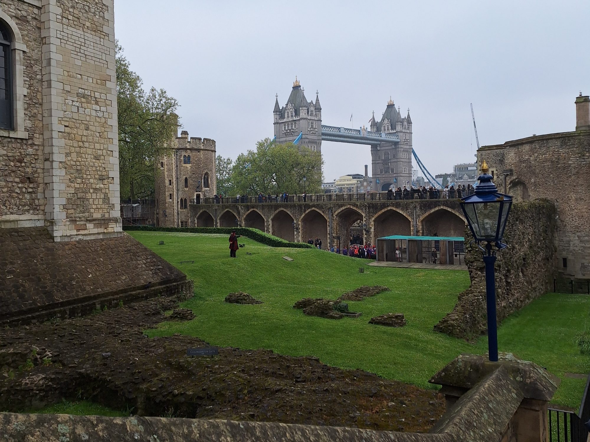 Inside the Tower of London with stone walls, green lawn, and Tower Bridge in the background.